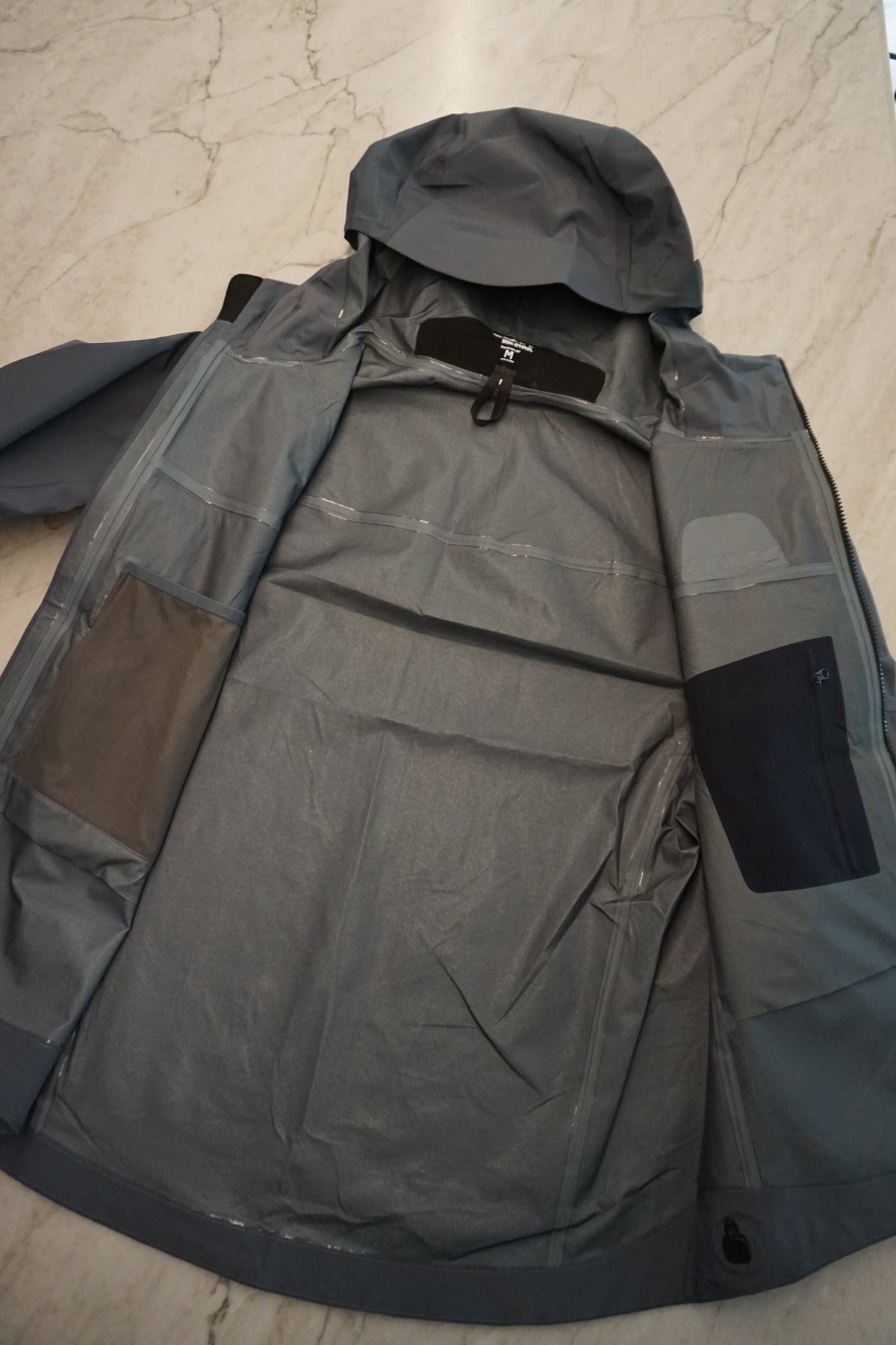Open grey technical shell on a marble background showing the internal drop pocket and security pockets, along with the micro seam taping.
