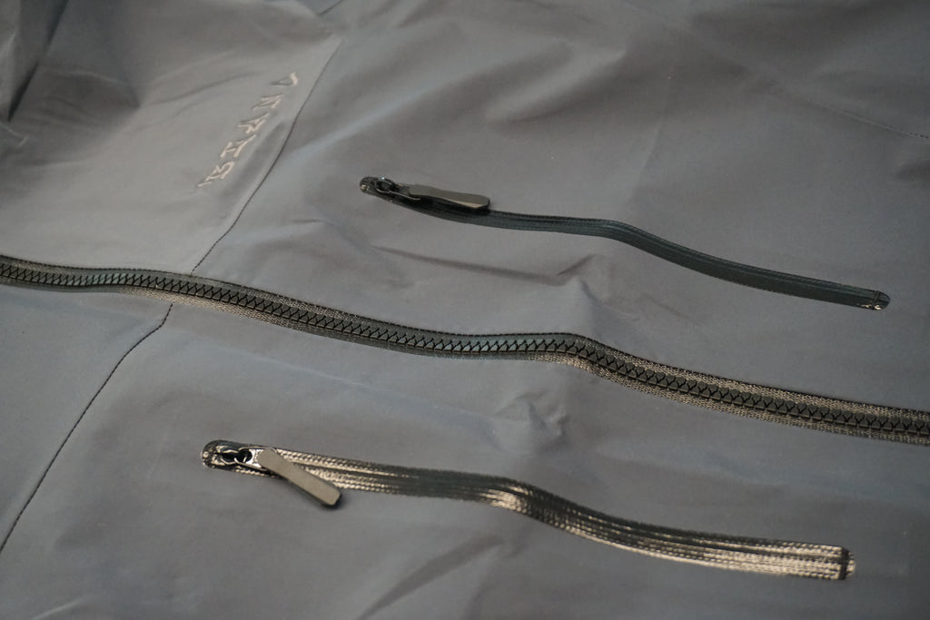 Grey technical shell with black chest mounted zippers and a tone on tone embroidered logo on the left side of the chest.