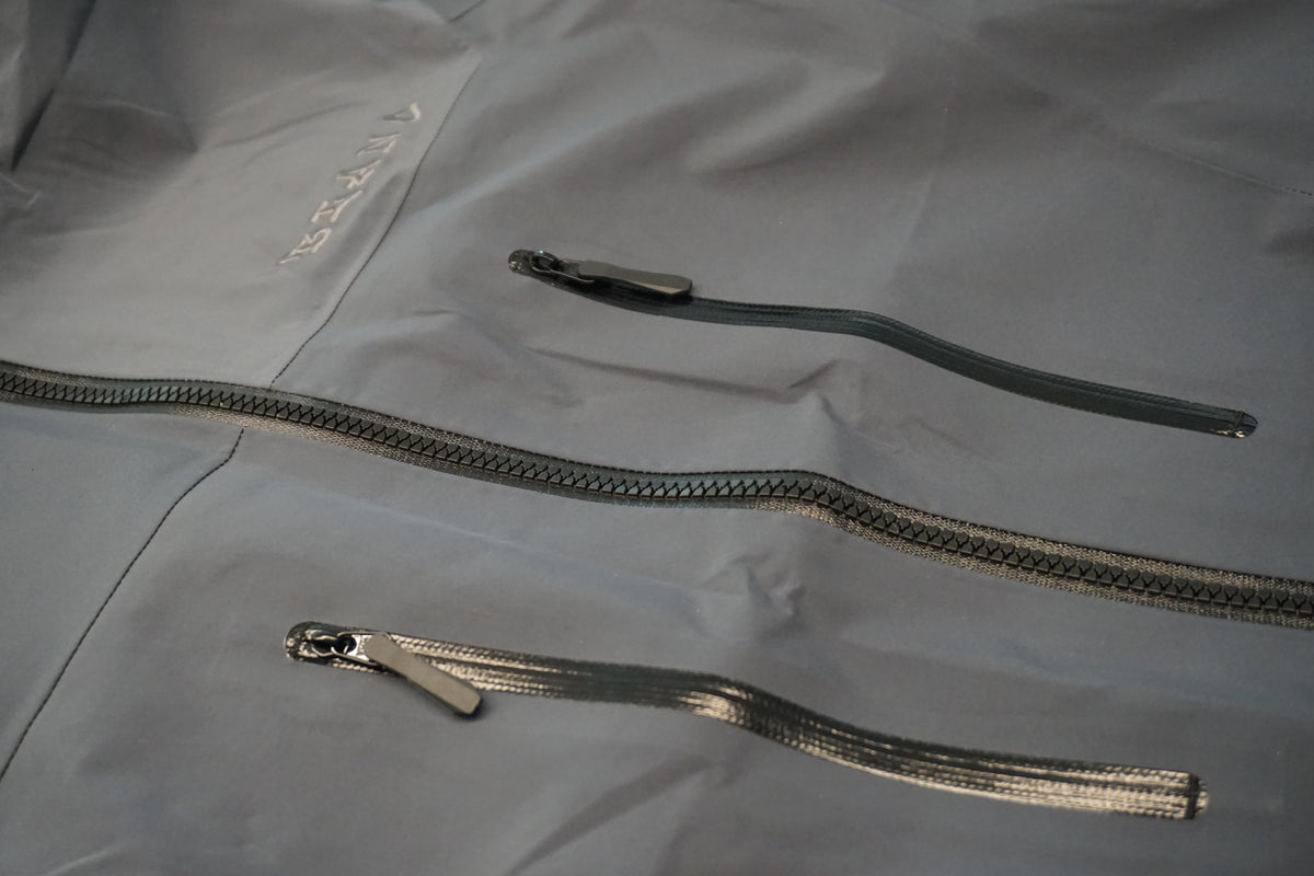 Grey technical shell with black chest mounted zippers and a tone on tone embroidered logo on the left side of the chest.