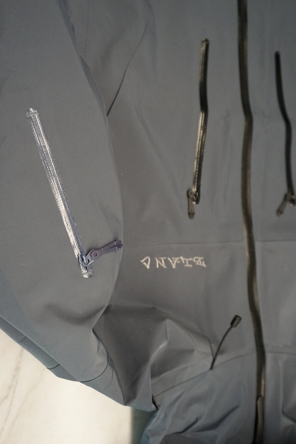 Close-up of a gray technical jacket with multiple zippered pockets and a small tone on tone embroidered logo on the chest.