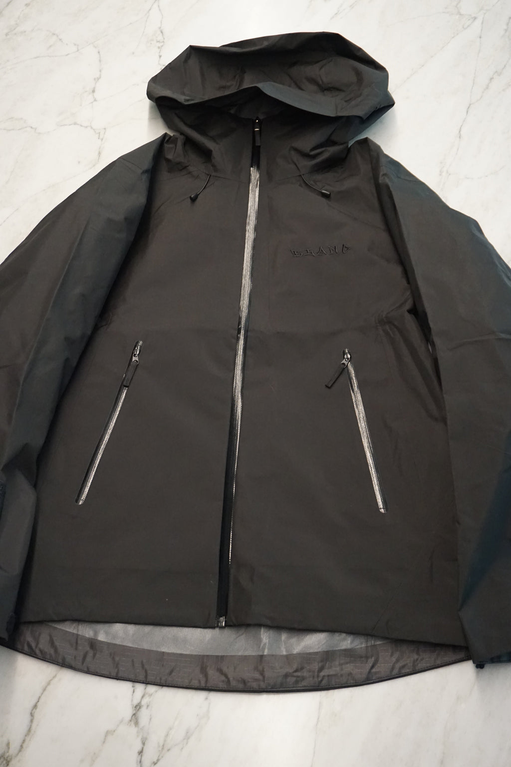 A black hooded jacket laid flat on a light marble surface, featuring a full front zipper and two diagonal zippered pockets.