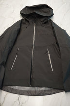 A black hooded jacket laid flat on a light marble surface, featuring a full front zipper and two diagonal zippered pockets.