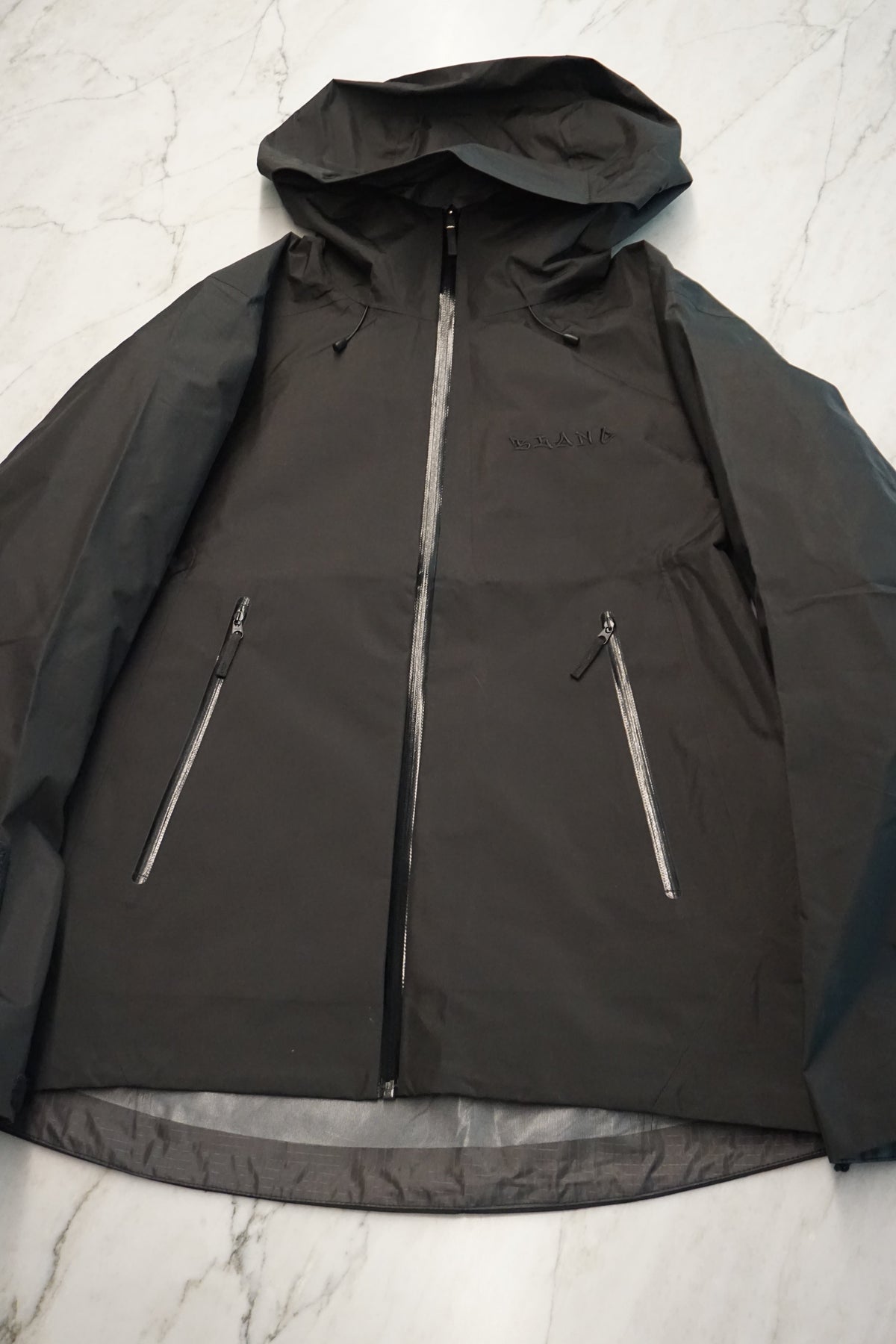 A black hooded jacket laid flat on a light marble surface, featuring a full front zipper and two diagonal zippered pockets.