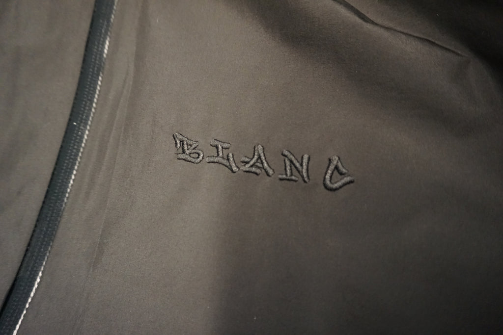 Close-up of a black jacket with a tone on tone embroidered 'BLANC' logo on the chest and a dark zipper running along the left side.