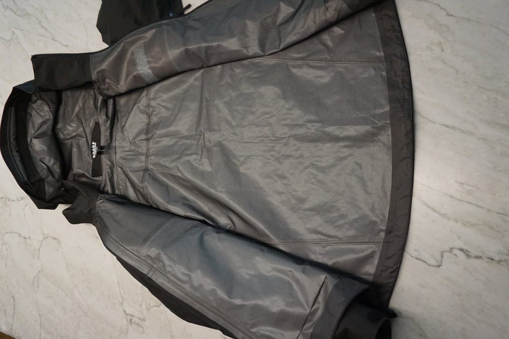 A black waterproof jacket with black trim and a hood, laid flat on a light gray marble surface.