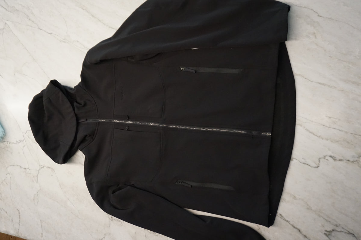 A black hooded softshell jacket laid flat on a white marble-patterned surface, featuring a full front zipper, zippered chest pockets, and a relaxed fit.