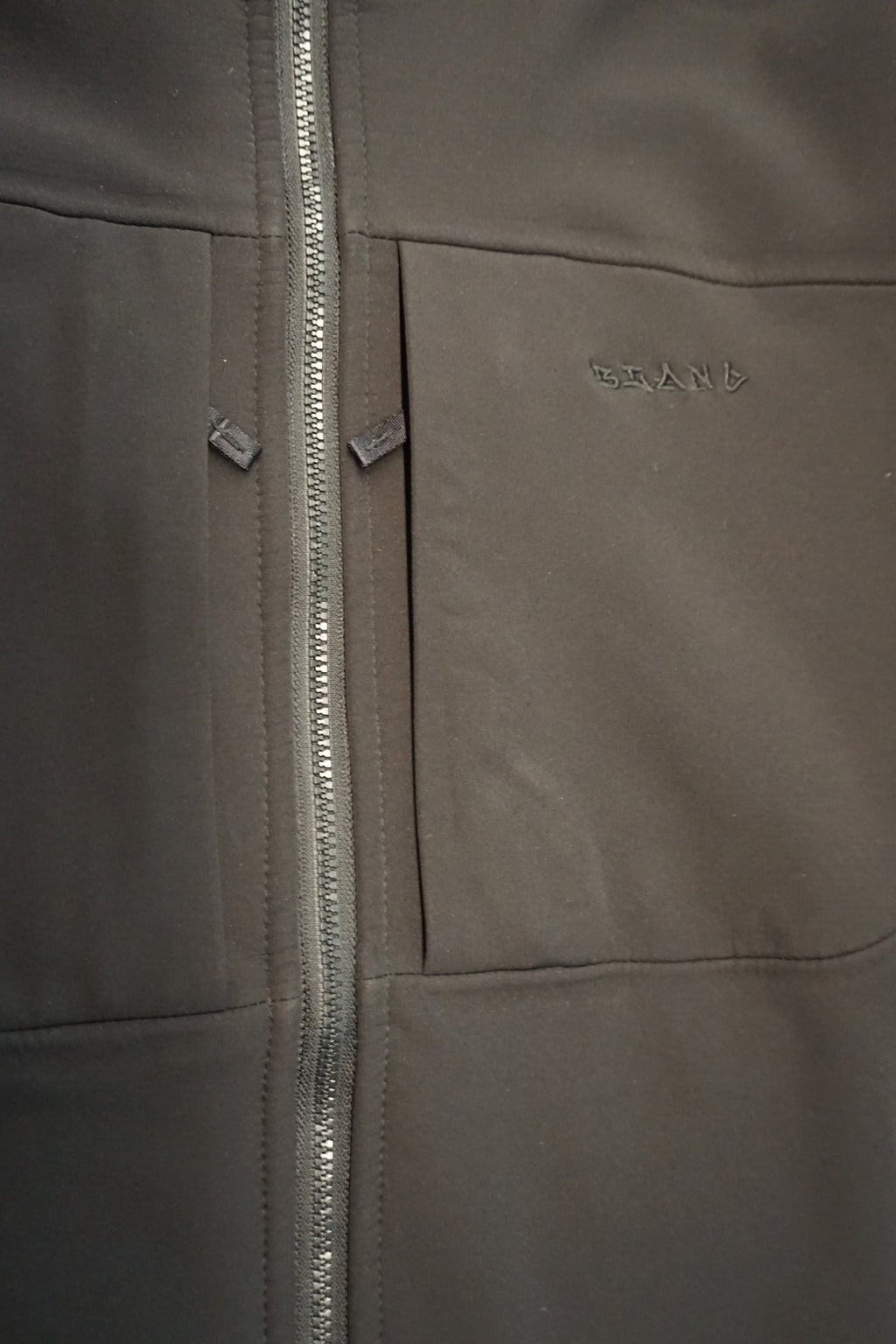 Close-up of a black softshell jacket showing a YKK Vislon zipper, 2 zippered pocket with an embroidered brand name 'Blanc' on the chest.