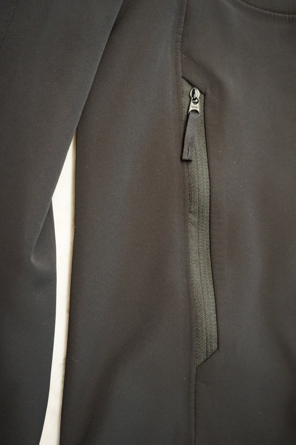 Close-up of a blacksoftshell jacket featuring a vertical zippered pocket with a textured fabric insert and a black YKK zipper pull.