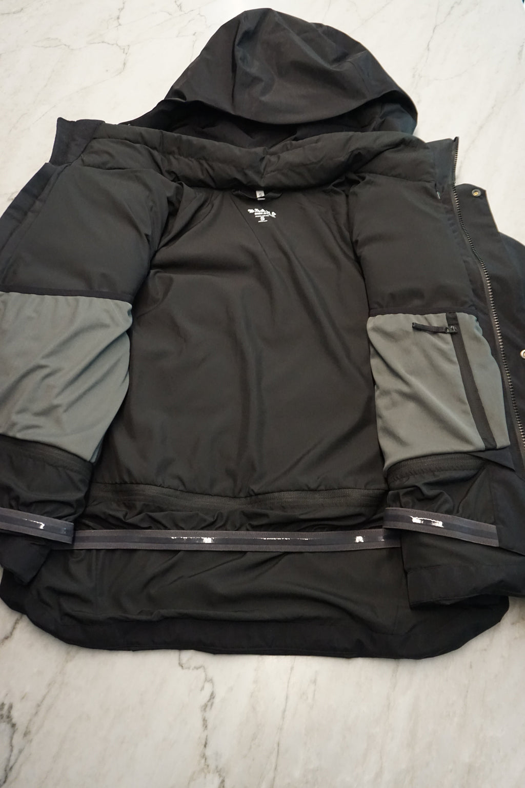 A black technical jacket laid flat and open on a light marble surface, showing the internal pockets and detachable powder skirt.