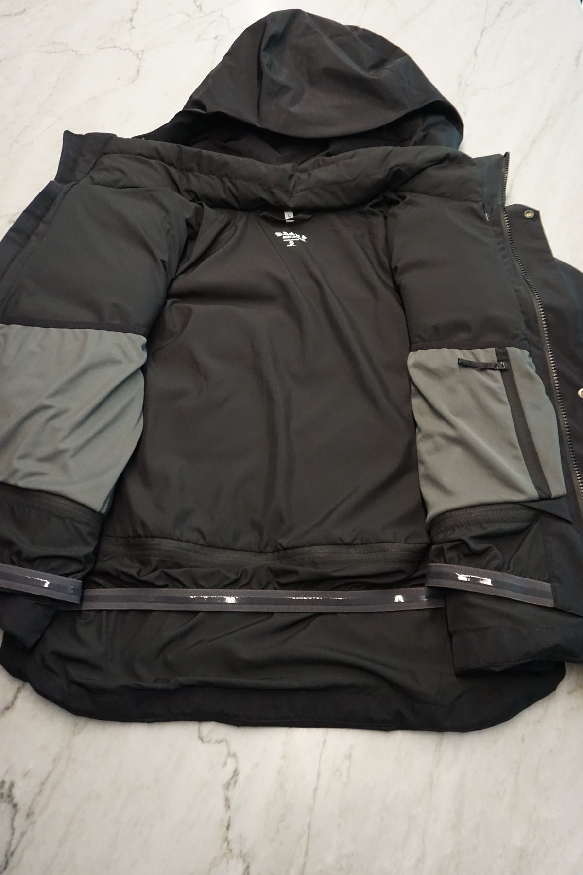 A black technical jacket laid flat and open on a light marble surface, showing the internal pockets and detachable powder skirt.