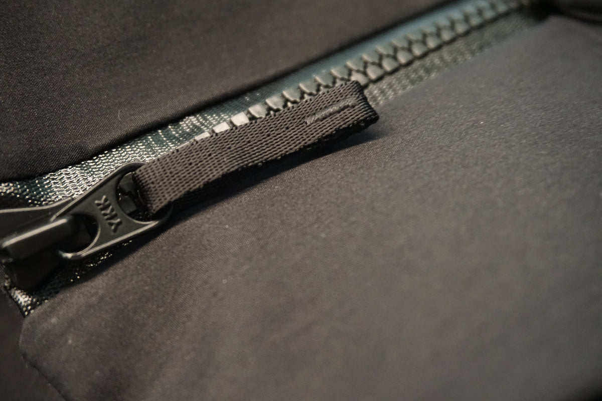 Close-up of a black zipper pull tab with a textured fabric loop and a black metal YKK aquaguard 2 zipper head zipper.