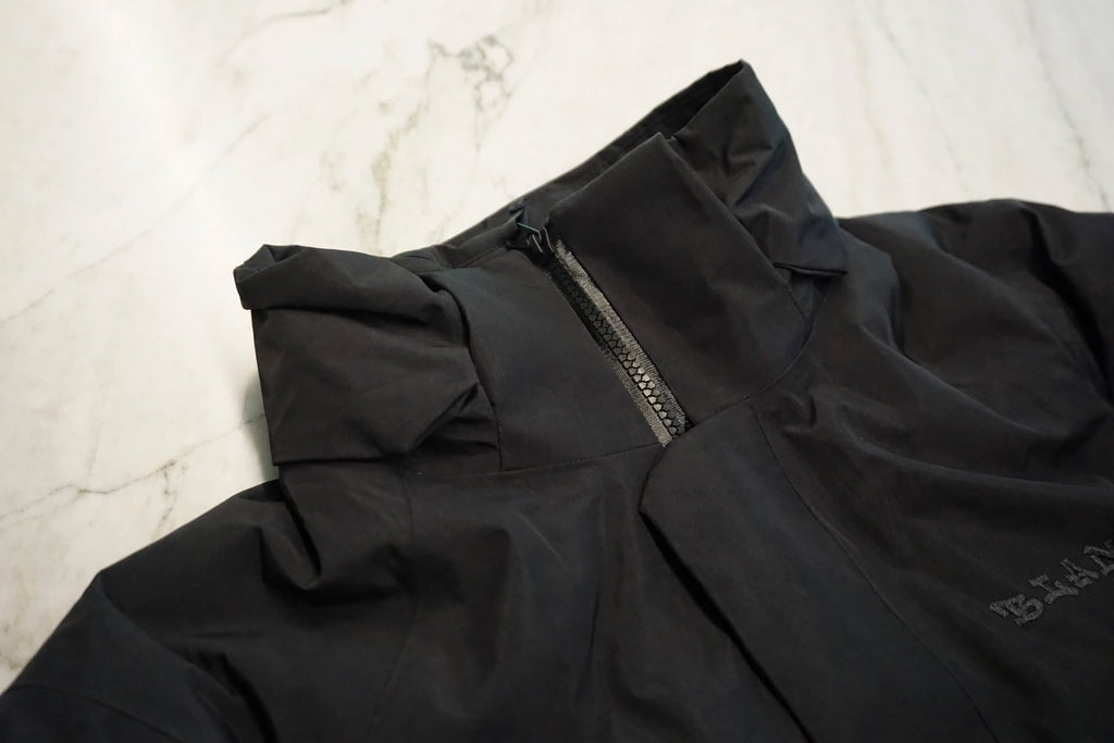 Close-up of a black waterproof jacket with a high collar and zipper, laid flat on a light marble surface. A small white logo is visible on the lower right side.