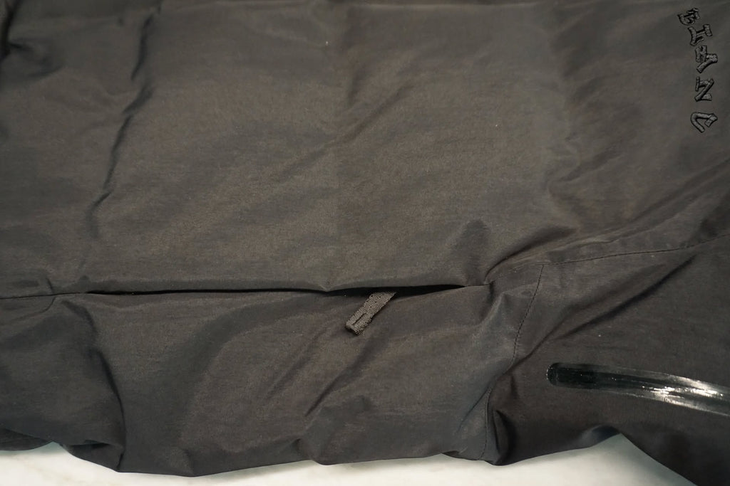 Close-up of a black waterproof jacket, showing a horizontal seam, a zipper pocket, and a tone on tone embroidered logo reading 'BLANC' on the upper right.