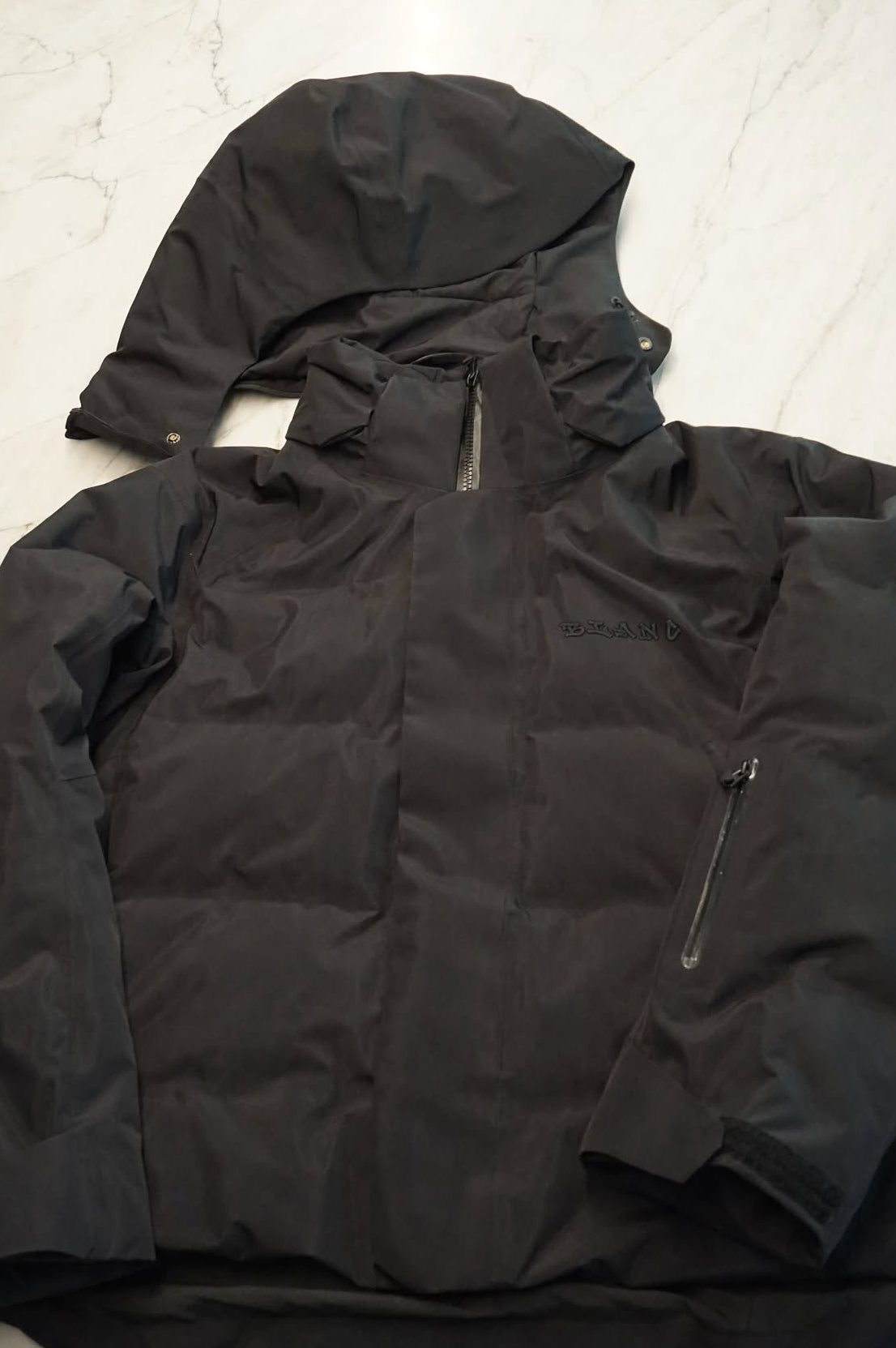 A black hooded puffer snow jacket laid flat on a light marble-patterned surface, featuring a front zipper, zippered chest pocket, and adjustable cuffs.